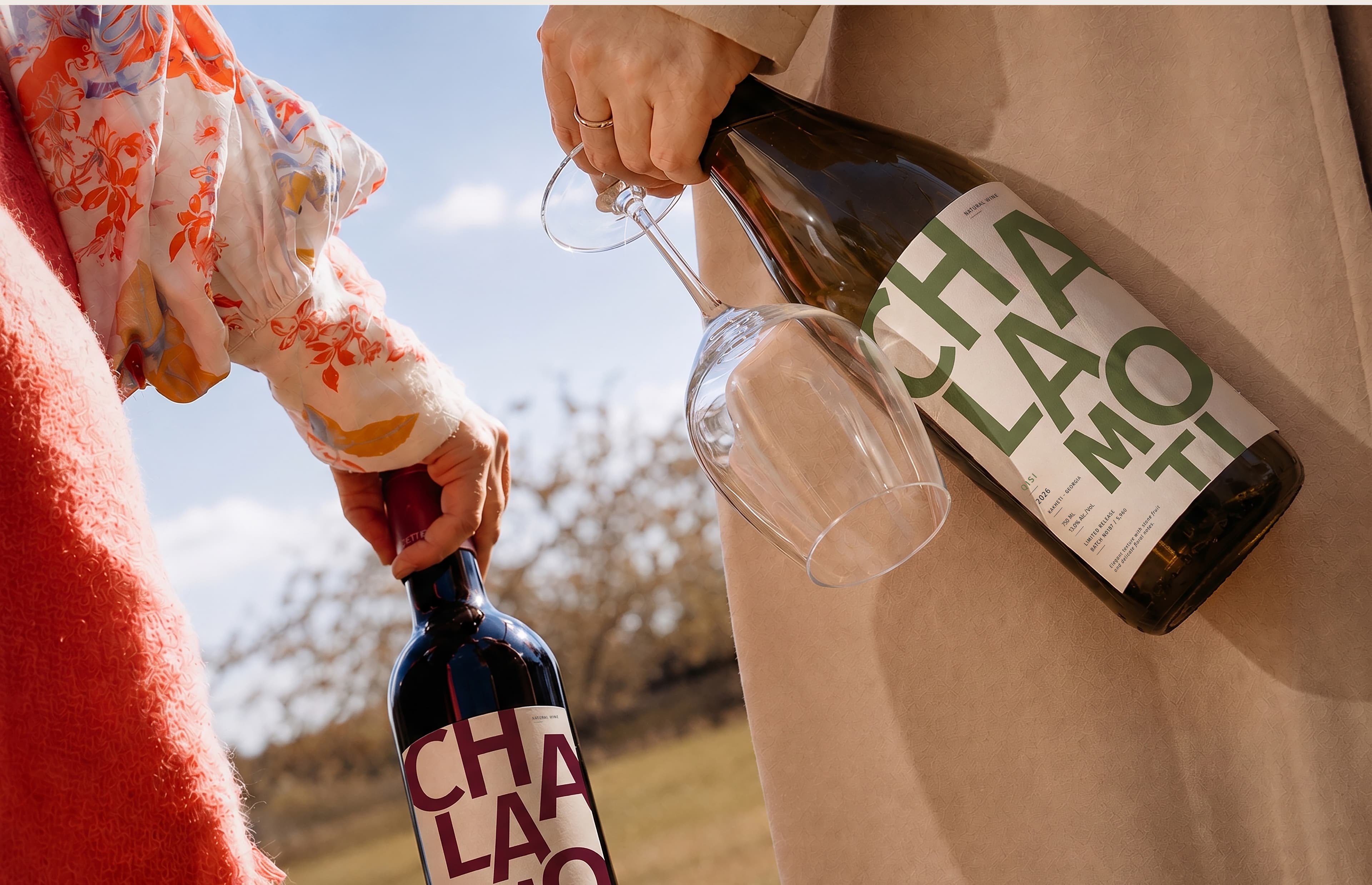 Two people carrying CHALAMOTI wine bottles and glasses outdoors.